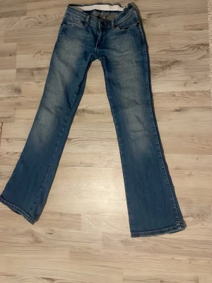 Blå bootcut jeans från Wrangler - Säljer ett par klassiska blå jeans från Wrangler med bootcut passform. snygga slitningar och ett brunt Wrangler-märke på bakfickan. Tillverkade i ett tåligt dimentyg.