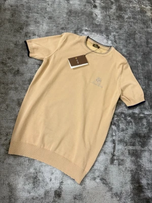 Beige t-shirt från Loro Piana - Snygg beige t-shirt från Loro Piana med broderad logga på bröstet. T-shirten har ribbade muddar och mörkblå detaljer vid krage och ärmslut. Tillverkad i mjuk bomull för en clean och exklusiv look.