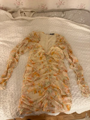 Blommig kortklänning från Gina Tricot - Supersöt kortklänning från Gina Tricot i ljusa toner med blommigt mönster i gult, orange och grönt. Klänningen har långa puffiga ärmar, v-ringning och snygga rynkade detaljer framtill. Tillverkad i ett lätt och luftigt polyestertyg, perfekt för sommarens alla tillfällen. Använd en gång❣️