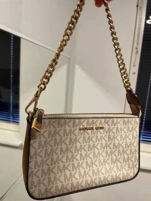 Beige axelväska från Michael Kors - Snygg beige axelväska från Michael Kors med kedjehandtag i guld och klassiskt MK-mönster. Väskan har bruna skinnsidor och dragkedja upptill. Insidan är fodrad med logotypmönstrat tyg och har en innerficka. Perfekt storlek för det viktigaste.