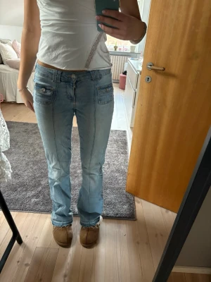 Ljusblå bootcut jeans med fickdetaljer - Säljer ett par ljusblå jeans med bootcut passform och snygga fickor framtill med knappdetaljer. Byxorna har låg midja och klassisk jeanslook med sömmar längs benen. Perfekta för dig som gillar retrovibbar och vill ha något som sticker ut lite extra.