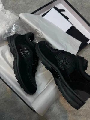 Svarta sneakers från Chanel - Säljer ett par svarta Chanel sneakers med chunky sula och ikoniska CC-loggan på sidan. Skorna är i en mix av mocka och skinn med snygga ton-i-ton detaljer och snörning framtill. Perfekta för dig som vill ha en clean och lyxig vibe till din outfit. Size 42-43