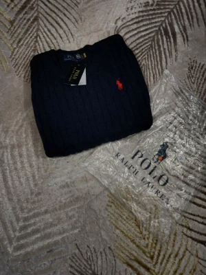 Marinblå stickad tröja Polo Ralph Lauren - Snygg marinblå stickad tröja från Polo Ralph Lauren med klassisk kabelstickning och röd broderad logga på bröstet. Tröjan har rund hals och ribbade muddar. Perfekt för dig som vill ha en stilren och tidlös look.