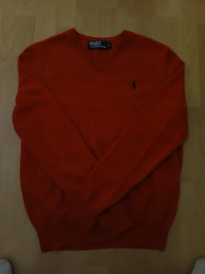 Röd v-ringad tröja Polo Ralph Lauren. - Säljer en röd v-ringad tröja från Polo Ralph Lauren med klassisk broderad logga på bröstet. Tröjan har lång ärm och är tillverkad i mjuk ull som känns skön mot huden. Perfekt för dig som gillar stilrena och tidlösa plagg. Bredd: 43cm / Längd: 63cm. Nypris: 725kr / Mitt pris: 290kr