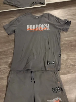 Grå Hoodrich t shirts & shorts dress  - Säljer ett par gråa Hoodrich shorts i storlek L. De har broderad logga i vitt och orange framtill, patch med text och coola orangea sömdetaljer. Shortsen har snörning i midjan och är gjorda i mjuk bomull, perfekta för streetwear och chill.          L på t shirten för de var liten i storlek                             M på shortsen 