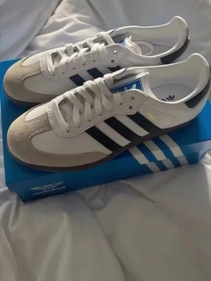 Adidas Samba OG vita och svarta sneakers - Adidas Samba OG sneakers i vitt och svart med klassiska tre ränder på sidan. Säljer pga jag fick de som present och jag tycker inte om skorna. De är helt nya aldrig använda.