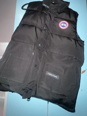 Svart dunväst från Canada Goose - Säljer en svart dunväst från Canada Goose med klassisk logga på bröstet. Västen har stora framfickor, hög krage och stängs med både dragkedja och tryckknappar. Perfekt för lager-på-lager och riktigt snygg till höst och vinter.
