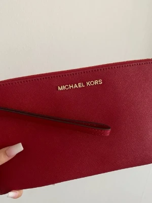 Röd clutch från Michael Kors - Snygg röd clutch från Michael Kors med guldfärgad dragkedja och logga framtill. Inga skador - i nyskick!