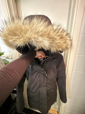 Svart parkas från Canada Goose - Säljer en svart Canada Goose parkas med stor huva och fluffig pälsdetalj. Jackan har flera praktiska fickor, dragkedja och knappar framtill samt den klassiska röda loggan på ärmen. Perfekt för kalla vinterdagar och riktigt snygg stil. Jackan är i strl XS, men är som i strl S/M