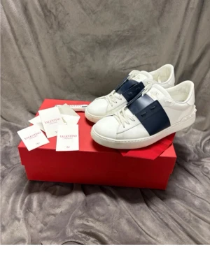 Valentino Garavani Open sneakers vit/blå - Säljer ett par Valentino Garavani Open sneakers i vitt skinn med bred marinblå rem över vristen och ikoniska nitar på hälen. Klassisk rund tå, vita snören och platt sula. Skorna är i otroligt bra skick och endast använda ett fåtal gånger. Kommer med originalkartong och tags.