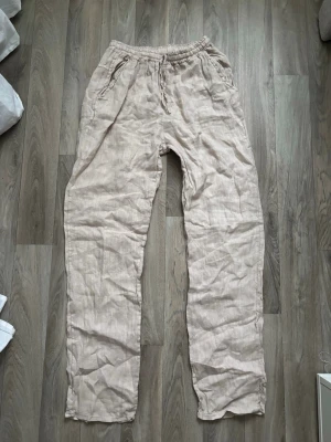 Beiga linnebyxor  - Säljer ett par beiga linnebyxor från Design by si med avslappnad passform och resår i midjan. Byxorna har snörning i midjan och två sidofickor. Perfekta för varma dagar.