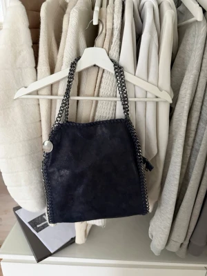 Mörkblå axelväska med kedjedetaljer - Säljer en mörkblå axelväska i syntetmaterial med snygga silverfärgade kedjor som axelband och runt kanterna. Väskan har dragkedja upptill och en extra ficka med dragkedja på baksidan. Perfekt storlek för det viktigaste och ger en cool vibe till din outfit.