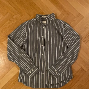 Randig blå skjorta från GANT - Säljer en klassisk blå skjorta från GANT med vita vertikala ränder. Skjortan har regular fit, lång ärm, knappar framtill och bröstficka. Tillverkad i 100% bomull för en skön känsla och tidlös look.