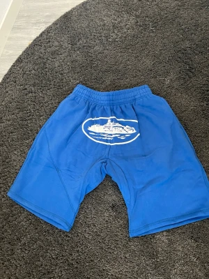 Corteiz Blå shorts med tryck - Säljer ett par blå Corteiz shorts med resår i midjan och ett vitt tryck framtill som föreställer ett fartyg. Shortsen har en avslappnad passform och är perfekta för sommardagar eller träning. Materialet känns mjukt och bekvämt. Storlek M men väldigt baggy fit så skulle säga det passar mer L. Hör av dig vid funderingar.