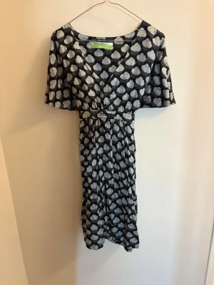 Svart klänning med molnmönster från Smirk - Unik svart klänning från Smirk med vitt molnmönster över hela tyget. Klänningen har v-ringning, korta ärmar och ett knytband i midjan för en snygg siluett. Materialet känns mjukt och lätt, perfekt för en cool och avslappnad look.