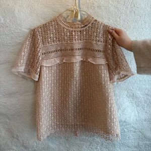 Beige spetsblus med volangdetalj - Säljer en superfin beige blus i spets med korta ärmar och hög hals. Blusen har ett genombrutet mönster, volangdetalj framtill och transparenta partier. Perfekt för dig som gillar romantisk och feminin stil.