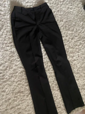 Svarta kostymbyxor  - Snygga svarta kostymbyxor med klassisk rak passform och pressveck. Byxorna har bälteshällor, fickor bak och är tillverkade i ett slätt tyg som ger en stilren look. Perfekta för dig som gillar en clean och dressad stil.