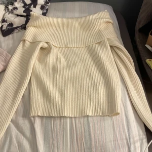 Ribbad offshoulder stickad tröja - Säljer en ljusbeige stickad tröja med ribbad struktur och offshoulder-design. Tröjan har långa ärmar och en bred krage som vilar snyggt över axlarna. Perfekt för dig som gillar en chill och trendig look med lite extra edge.