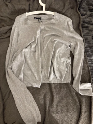 Grå kofta med knappar - Snygg grå kofta från  med rund halsringning och knappar hela vägen framtill. Långärmad modell i mjukt och tunt stickat material, perfekt att slänga på sig när det är lite kyligt. Enkel och stilren design som funkar till många olika looks.