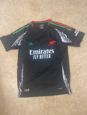 Arsenal x Adidas svart matchtröja - Svart Arsenal matchtröja från Adidas med gröna och röda detaljer på axlarna, vitt tryck på bröstet och klubbens logga i rött. Tröjan har grafiska mönster längs sidorna och är tillverkad i lätt polyester med Aeroready-teknologi.