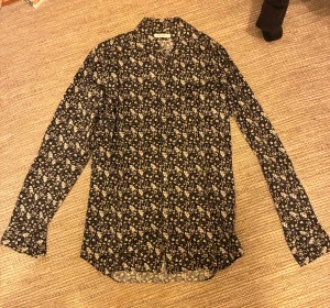 Blommig svart skjorta från J.Lindeberg - Säljer en blommig skjorta från J.Lindeberg med snyggt blommigt mönster i vitt. Skjortan har klassisk krage, knäppning framtill och långa ärmar. Perfekt för dig som vill sticka ut lite extra i sommar på golfbanan eller på sommarfester. Nypris 999