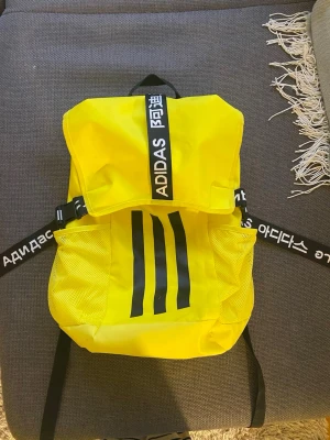 Adidas ryggsäck - Gul sportig ryggsäck från Adidas med svarta detaljer och logga på remmarna. Väskan har tre svarta ränder på framsidan, meshfickor på sidorna och justerbara axelremmar. Tillverkad i 100% återvunnen polyester