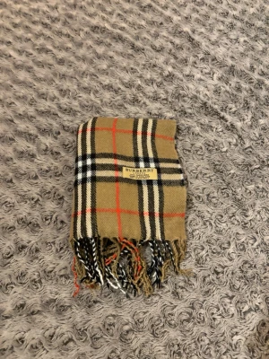 Burberry halsduk - Säljer en klassisk rutig halsduk från Burberry i 100% kashmir. Färgerna är beige, svart, vit och röd med fransar i ändarna. Den har den ikoniska Burberry-taggen och är tillverkad i England. Perfekt accessoar för att lyfta din look. Den är äkta.