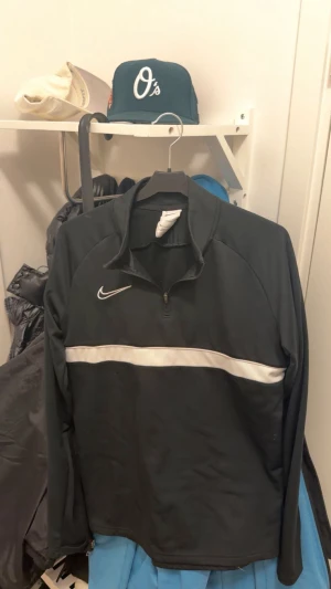 Svart Nike half zip tröja - Svart långärmad Nike-tröja med vit rand över bröstet och klassisk swoosh-logga. Tröjan har half zip-dragkedja och sportig vibe, perfekt för träning eller chill. Tillverkad i mjukt syntetmaterial som andas.