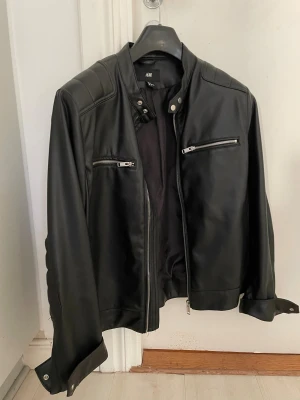 Svart skinnjacka från H&M - Ny Snygg svart skinnjacka från H&M med coola quiltade detaljer på axlar och ärmar. Jackan har två dragkedjefickor fram, ståkrage med tryckknappar och dragkedja framtill. Perfekt för dig som gillar en edgy stil.endast provad 