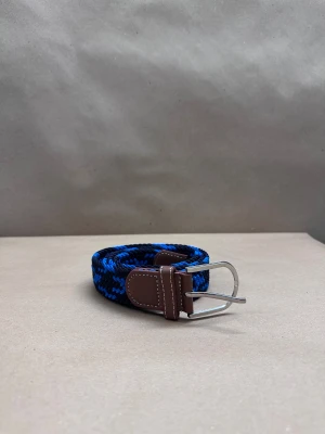 Blå och svart flätad bälte - Snyggt flätat bälte i blått och svart med bruna detaljer i skinn och silvrigt spänne. Bältet har ett coolt mönster och är perfekt för att ge din outfit lite extra edge. Passar till jeans eller chinos och är lätt att justera tack vare den flexibla designen.