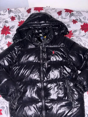 Svart pufferjacka från Polo Ralph Lauren - Säljer en svart, glansig pufferjacka från Polo Ralph Lauren med huva och dragkedja framtill. Jackan har det klassiska röda Polo-logot broderat på bröstet, två sidofickor med dragkedja och elastiska muddar vid ärmsluten. Perfekt för kalla dagar.