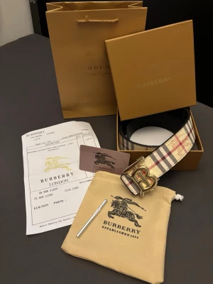 Burberry rutig skinnväst med guldspänne - Snygg Burberry väst i klassiskt rutigt mönster med beige, svart och röd ton. Västen har ett stort guldigt 'B'-spänne framtill och är tillverkad i skinn. Perfekt för dig som vill sticka ut med en ikonisk designerlook.