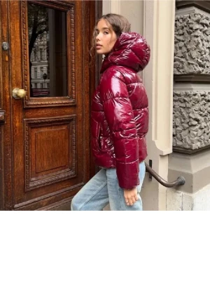 Vinröd pufferjacka med huva - Säljer en vinröd pufferjacka med glansig finish och stor huva. Jackan har lång ärm, dragkedja framtill och breda quiltade sektioner som ger en cool och modern look. Perfekt för dig som vill ha en varm och snygg jacka med lite oversized känsla.
