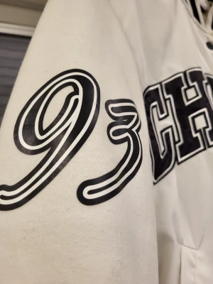 Vit bomberjacka college CHAMPS från H&M - Säljer en vit bomberjacka från H&M med svart-vita ribbade muddar och krage. Jackan har stora broderade bokstäver 'CHAMPS Series' på bröstet, tryckta detaljer som '93' på ärmen och 'The 1999 3rd Division' vid fickan. Stängs med svarta knappar och har två sidofickor.