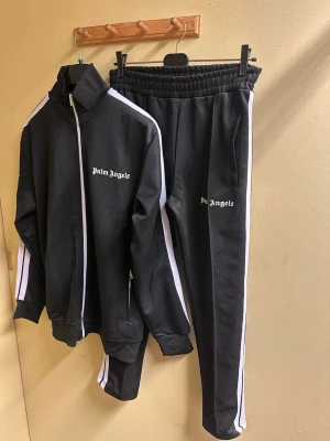 Palm Angels svart träningsset - Säljer ett svart träningsset från Palm Angels med vita ränder längs ärmar och ben. Jackan har hög krage, hel dragkedja och två fickor med dragkedja. Byxorna har resår i midjan och Palm Angels-logga på bröst och lår. Perfekt för en sportig och clean look.