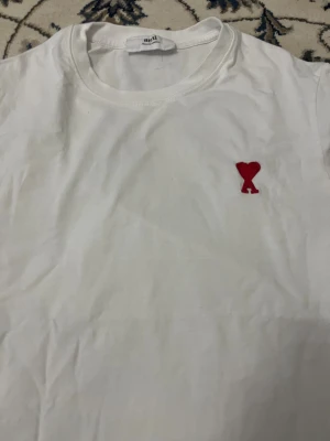 Vit t-shirt från Ami med rött hjärta - Vit t-shirt från Ami Alexandre Mattiussi med klassisk rund hals och korta ärmar. På bröstet finns ett broderat rött hjärta med A. Tillverkad i mjuk bomull för skön känsla. Perfekt för dig som gillar stilrena plagg med en twist.