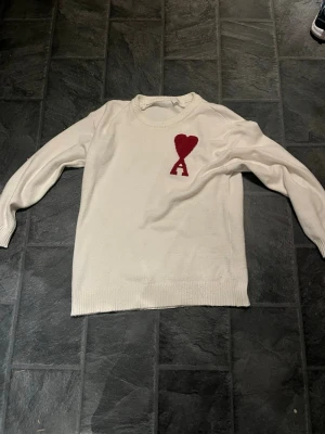 Vit stickad tröja från Ami Paris - Säljer en vit stickad tröja från Ami Paris med röd hjärtapplikation och bokstaven A på bröstet. Tröjan har rund hals och långa ärmar med ribbade muddar. Perfekt för dig som gillar stilrena detaljer och vill ha något som sticker ut. Har bara använts 2 gånger.passa på att köpa🌟