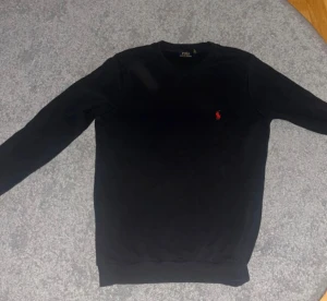 Svart tröja från Polo Ralph Lauren - Svart långärmad tröja från Polo Ralph Lauren med klassisk rund hals och röd broderad logga på bröstet. Tröjan har en enkel och stilren design, perfekt för dig som gillar tidlösa plagg med en touch av lyx.