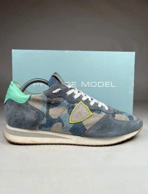 Philippe Model blå camo sneakers - Snygga sneakers från Philippe Model med blått och grått camouflage-mönster, detaljer i mocka och textil, vit snörning och silverfärgad plös. Bakkappan är mintgrön och sidologgan har en gul kant. Platt sula och rund tå för skön komfort.