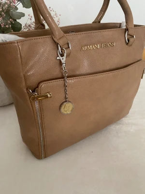 Beige handväska från Armani Jeans - Snygg beige handväska från Armani Jeans i skinn med guldfärgade detaljer och logotyp framtill. Väskan har två rejäla handtag, en praktisk ytterficka med dragkedja och en dekorativ nyckelring med AJ-emblem. Insidan är fodrad med mönstrat tyg med logotryck.