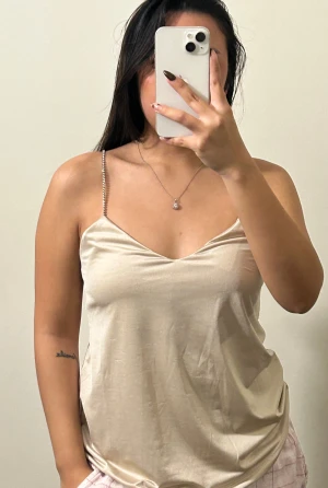 Champagnefärgad linnetopp med strass - Snygg champagnefärgad linnetopp med tunna axelband täckta av strass. Toppen har en lätt glansig finish och v-ringad halsringning. 