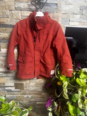Röd parkajacka Tretorn Sarek 122/128 - Parkajacka från Tretorn Sarek i en snygg röd färg. Jackan har hög krage, stora fickor med tryckknappar och dragkedja framtill. Fodrad med mjuk teddy på insidan för extra värme. Perfekt för kyliga dagar och outdoor vibes.