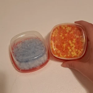 Två burkar med pärlor i blått, gult och orange - Säljer två små plastburkar med pärlor. Ena burken innehåller ljusblå pärlor och den andra har en mix av gula och orange pärlor. Perfekt soppa för pyssel eller smyckestillverkning. Burkarna har röda lock och är genomskinliga så man ser färgerna tydligt. 