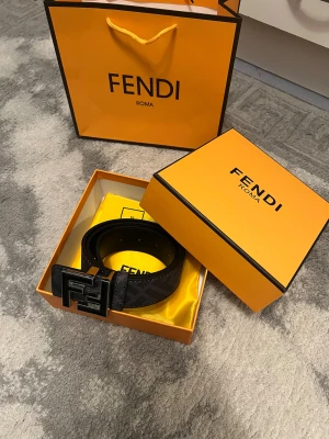 Svart Fendi bälte med FF-spänne - Säljer ett svart Fendi-bälte med ikoniskt FF-spänne i metall. Bältet har diskret rutigt mönster och är gjort i äkta skinn med präglad textur. Kommer i original ask och påse, perfekt för dig som vill ha en lyxig detalj till outfiten.