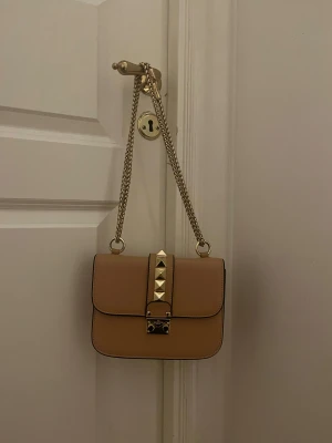Aprikos glam lock axelväska från Valentino - Snygg aprikos axelväska från Valentino med guldfärgade kedjor och coola pyramidnitar på framsidan. Väskan har ett stilrent, fyrkantigt format och är gjord i skinn. Perfekt för dig som gillar exklusiva detaljer och vill ha en väska som sticker ut. Modellen är glam lock vet ej om äktigheten därav priset jätte bra skick annars. 