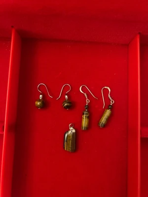 Brun och gul tigrisöga smyckeset - Säljer ett set med två par örhängen och ett hänge i tigrisöga. Örhängena har antingen rund eller avlång form och är i bruna och gula toner med metallfästen. Hänget är rektangulärt och har en metallögla. Perfekt för dig som gillar naturliga stenar och unika accessoarer.