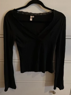 Svart omlottopp med spets från Nelly - Svart omlottopp från Nelly i storlek XS. Toppen har lång ärm, v-ringad hals med spetskant och en slim passform. Materialet är mjukt och stretchigt.