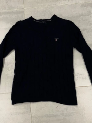 Mörkblå kabelstickad tröja GANT - Snygg mörkblå kabelstickad tröja från GANT i mjuk lammull. Klassisk rund hals och diskret broderad logga på bröstet. Tröjan har långa ärmar och ribbade muddar vid ärmslut och nederkant. Perfekt för dig som gillar stilrena och tidlösa plagg.