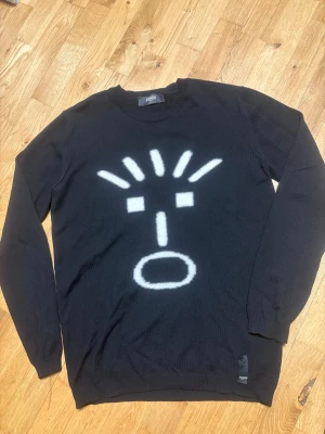 Svart Fendi ulltröja med grafiskt tryck - Svart stickad tröja från Fendi i 100% ull med ett stort vitt grafiskt ansikte tryckt på framsidan. Rund hals och långa ärmar, perfekt för dig som gillar statement-plagg och exklusiva märken. Italiensk design med mjuk och skön känsla. Använd max 5 ggr,  ny pris ca. 5000kr