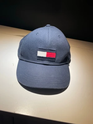Mörkblå keps från Tommy Hilfiger - Snygg mörkblå keps från Tommy Hilfiger med klassisk broderad logga i vitt, rött och blått framtill. Justerbar snapback baktill för perfekt passform. Tillverkad i bomull och har ventilerande hål upptill. Perfekt accessoar för en clean och sportig look.